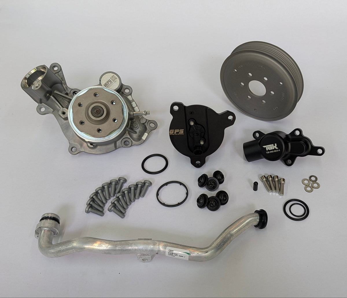 An automotive parts kit laid out on a white surface, including a silver metal pump, a black pulley, a black thermostat by German performance solutions, an aluminum radiator hose, gaskets, washers, and screws.